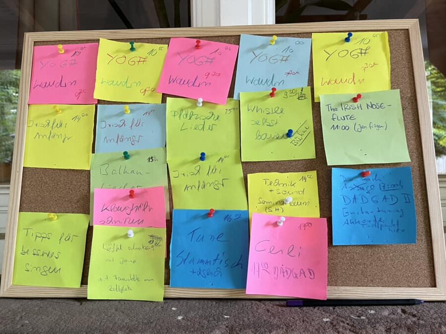 Das Bild zeigt die Pinnwand von der Sommer-Woche 2022: Auf einer Korktafel mit dünnem Holzrahmen sind 19 handschriftlich beschriftete Zettel in bunten Farben (gelb, pink, hellgrün, hellblau, mittelblau) gepinnt, jeweils in Spalten für die Wochentage. Zu lesen ist etwa "YOGA", "Wandern, "Ceili", "DADGAD", "Technik", "Balkan", "Irish Nose Flute", "Whistle" usw. und die Uhrzeiten dazu. 