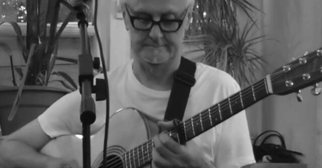 Das Schwarz-Weiß-Bild zeigt den Iain Goodwin beim Spiel an der Gitarre. Er trägt eine schwarze Brille und schaut konzentriert nach unten. Das Bild ist ein Screenshot aus einer Videoaufnahme von einem Event im Naturfreundehaus Elmstein im Rahmen der Folksounds.